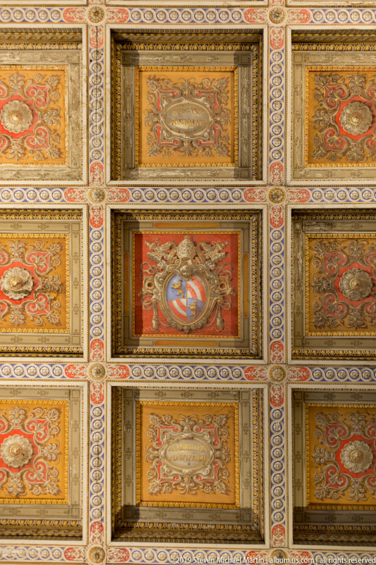 Close view of a red-and-gold coffered ceiling centered on a multicolored heraldic emblem within a rectangular panel.