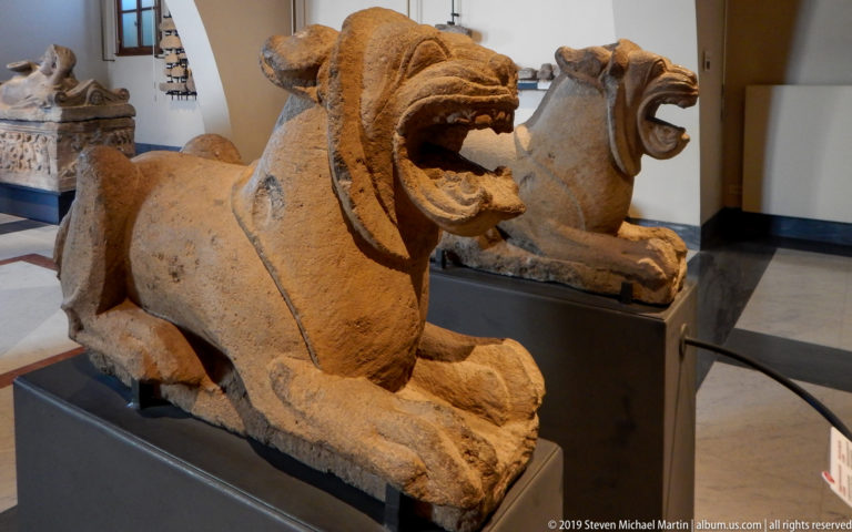 Weathered stone lion crouching with open jaws and deeply carved mane on a dark museum plinth, with another animal sculpture behind.