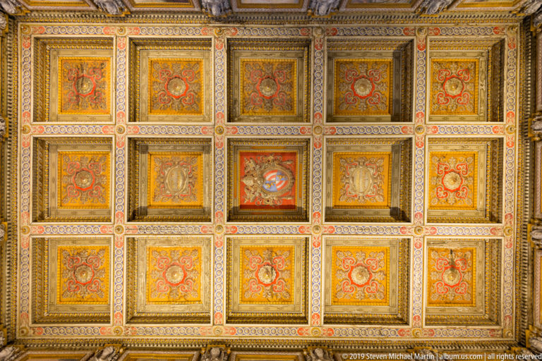 Rectangular grid of richly gilded ceiling coffers filled with red, gold, cream, and blue ornament and heraldic motifs.