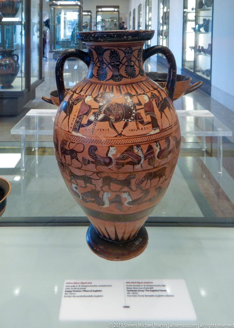 Large orange-and-black amphora with two handles, mythological figures, animal friezes, floral neck ornament, and a broad foot.