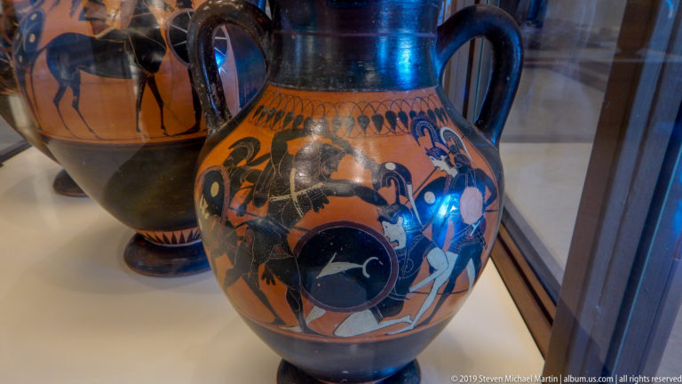 Tall orange-and-black amphora with several painted figures around a large circular shield motif, dark neck, and curved handles.