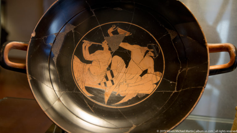 Shallow black ancient drinking cup with two side handles and an orange-red circular interior scene of several human figures.