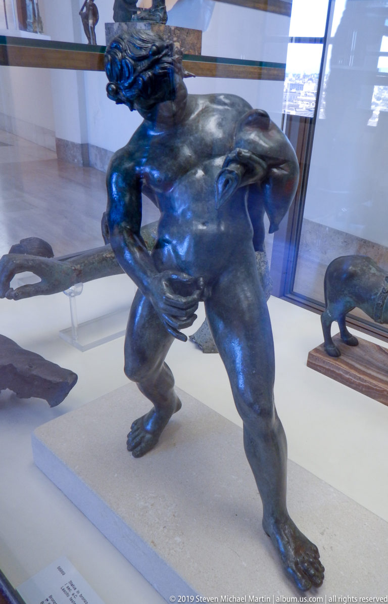 Dark bronze nude male figure striding while carrying another figure across his shoulders, displayed inside a museum case.