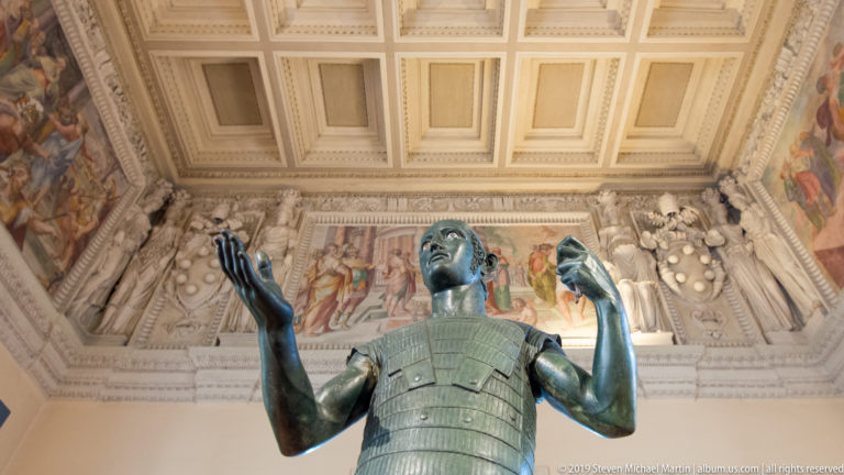 Low-angle view of a bronze standing male figure raising both hands beneath an ornate coffered museum ceiling.