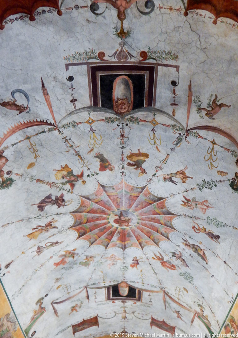 Close view of a painted ceiling centered on a multicolored starburst surrounded by figures, animals, and celestial motifs.