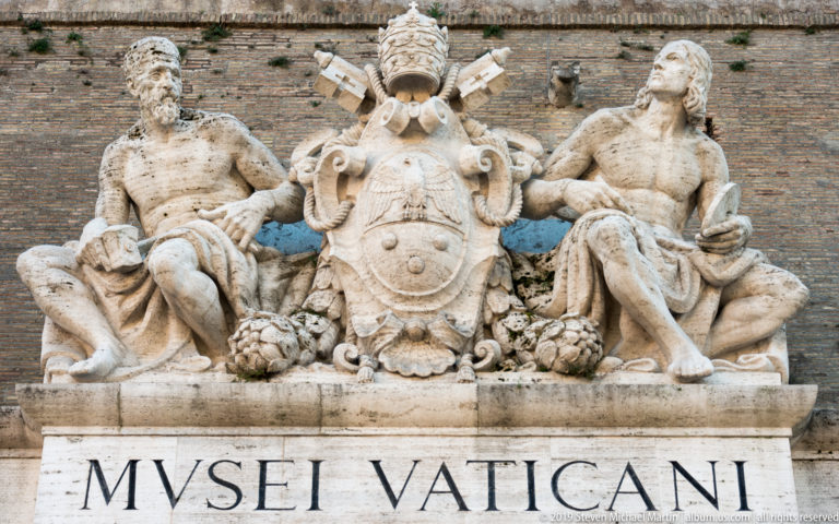 Close view of the Vatican Museums stone crest with reclining male figures, papal shield, tiara, crossed keys, and MUSEI VATICANI inscription.