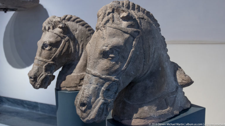 Two weathered carved stone horse heads on dark museum mounts, with visible bridles, nostrils, and stylized manes.