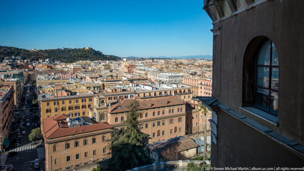 Elevated panorama of Rome's dense ocher and terracotta buildings, with a wooded hill at left and a museum window edge at right.