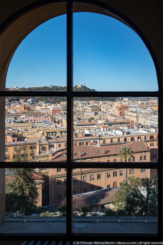Wide Rome panorama through a large arched window, with dense rooftops, Vatican buildings, trees, and distant hills.