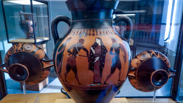 Large orange-and-black ancient amphora with painted figures and animals, displayed between two smaller decorated vessels.