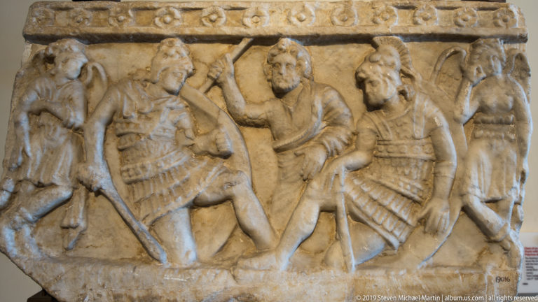 Weathered stone relief fragment with armored warriors, shields, raised weapons, and a central bearded figure in close combat.