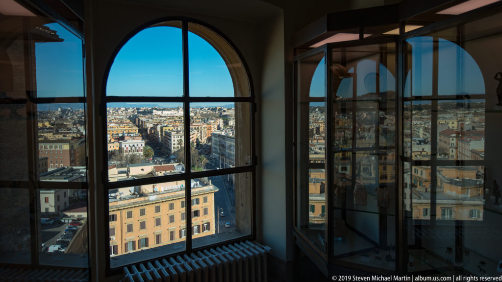 Rome rooftops and streets seen through tall arched Vatican Museums windows with dark mullions and interior reflections.