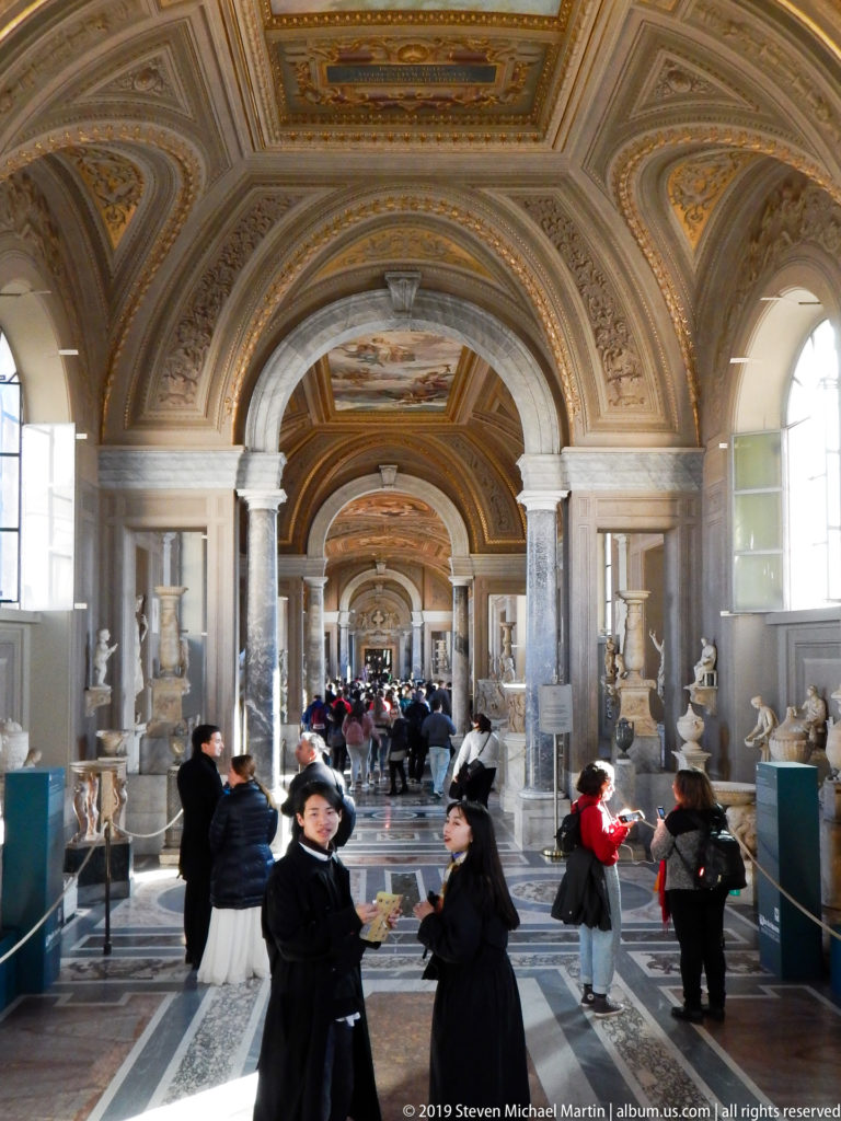 Long Vatican sculpture gallery with visitors, ancient statues, busts, urns, columns, and an ornate vaulted ceiling.