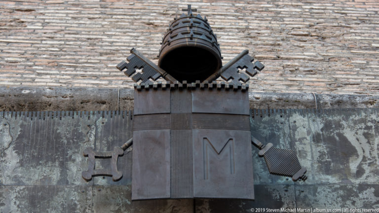 Dark metal papal emblem with tiara, crossed keys, and a rectangular panel marked M, mounted on a weathered masonry wall.