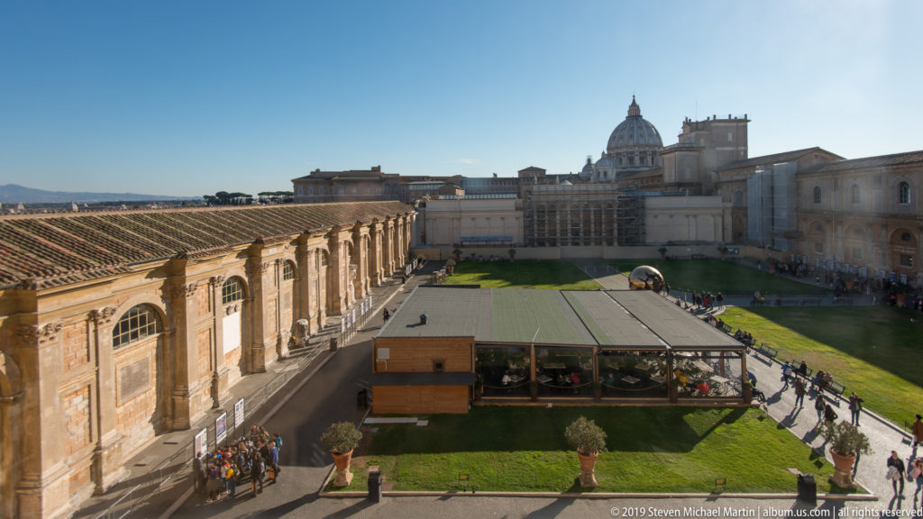 Elevated view across a Vatican Museums courtyard toward St. Peter's Basilica dome, with arched museum wings, lawns, and visitors.