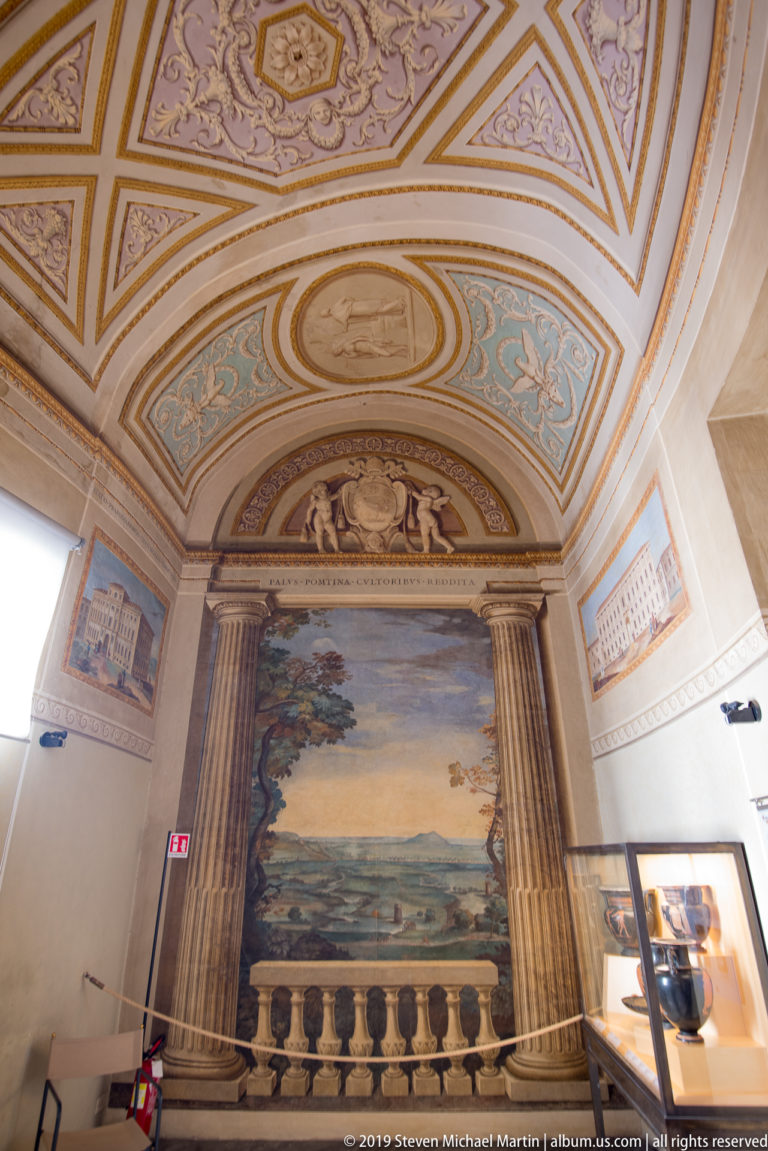 Museum landing with a large landscape painting, fluted pilasters, balustrade, and pastel pink-and-blue ornamental ceiling.