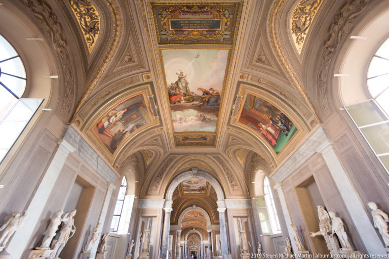 Long vaulted Vatican gallery with classical statues on both sides, large painted ceiling panels, arches, and high windows.