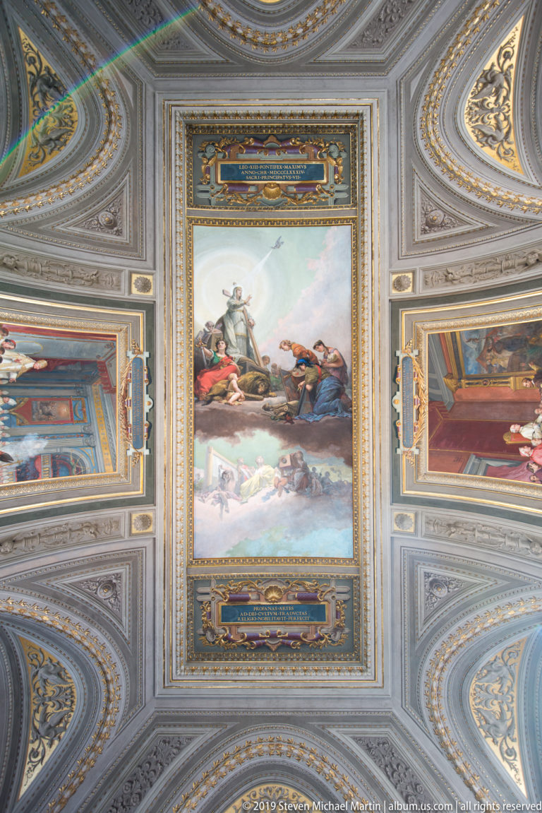 Rectangular battle or military painting centered in a pale-gray and gold decorated ceiling with smaller colored panels around it.