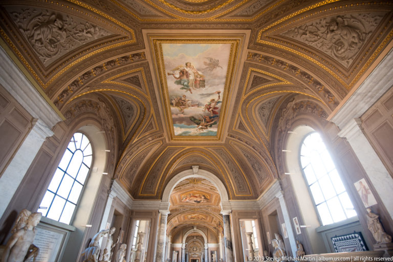Long Vatican Museums gallery with tall windows, sculptures, arches, and an ornate frescoed and gilded vaulted ceiling.