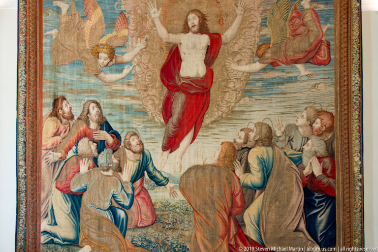 Tapestry of Christ ascending with raised arms above apostles and angels, his red drapery prominent against pale clouds.
