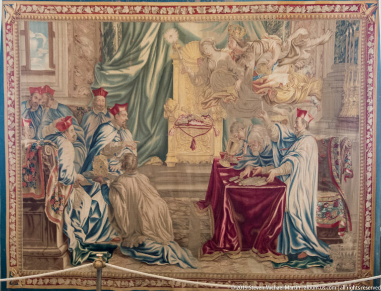 Large tapestry of richly robed clerics gathered around a table, with figures appearing in clouds above and an ornate woven border.