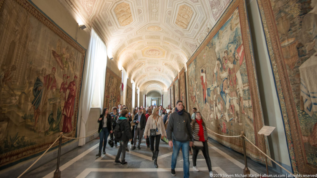 Visitors walking through a long Vatican gallery lined with large narrative tapestries beneath a pale decorated vaulted ceiling.