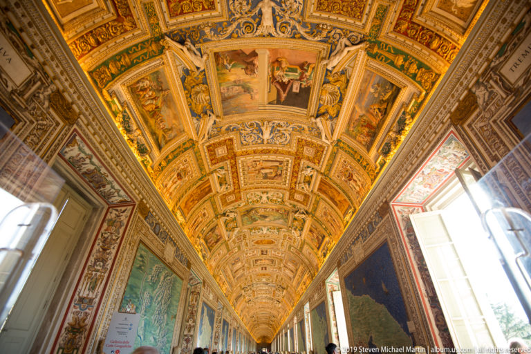 Long Gallery of Maps with painted maps on both walls, visitors, tall windows, and a richly gilded barrel-vaulted ceiling.