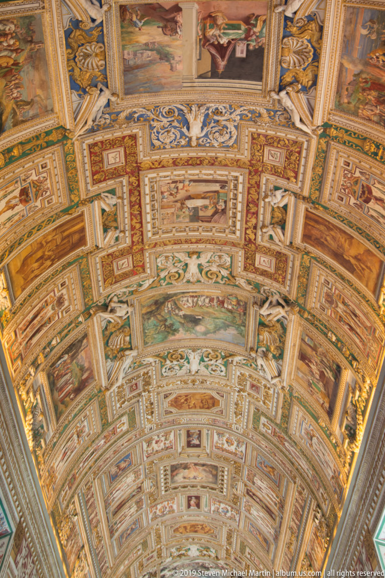 Upward view of an ornate barrel vault covered with painted scenes, geometric compartments, foliage, and gilded borders.