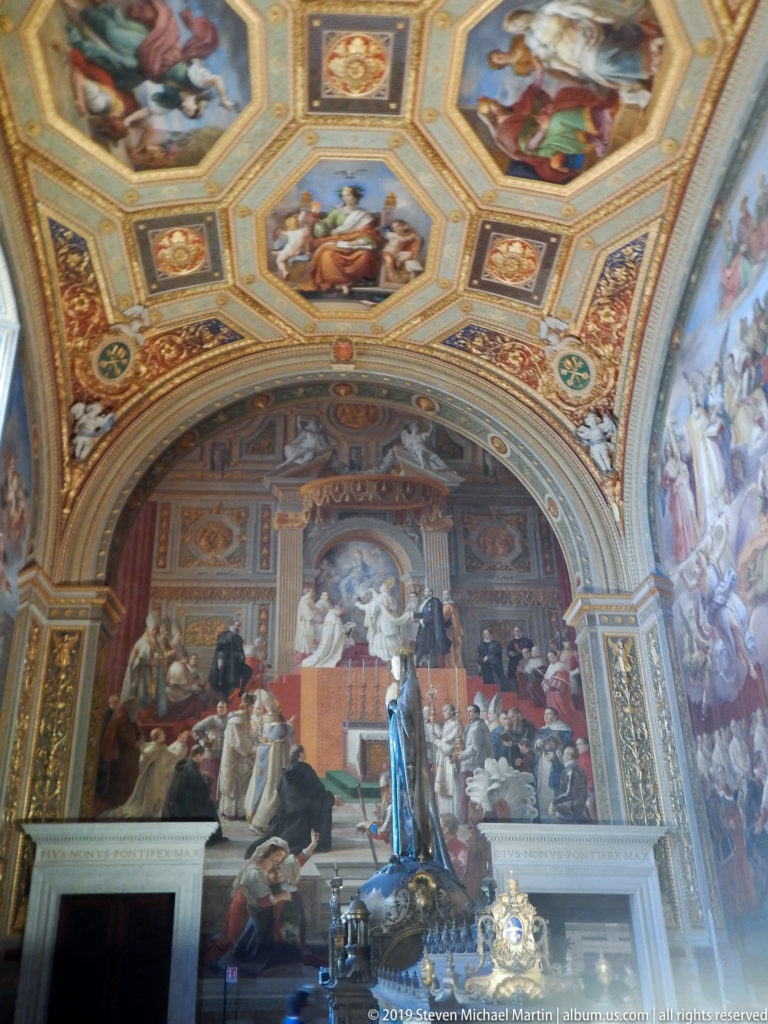 Wide Vatican room with a papal ceremonial fresco, dark foreground statue, gilded octagonal ceiling panels, and adjacent wall paintings.