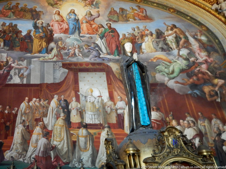 Papal ceremonial fresco with clergy below saints and sacred figures, partly obscured by a dark statue and ornate foreground metalwork.