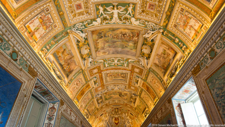 Long Gallery of Maps barrel vault covered with gilded frames, painted scenes, and dense ornament, viewed along the gallery axis.