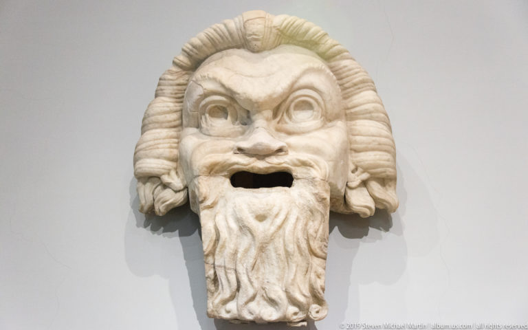 Carved marble theatrical mask with bulging eyes, arched brows, open mouth, thick hair, and a long curling beard.