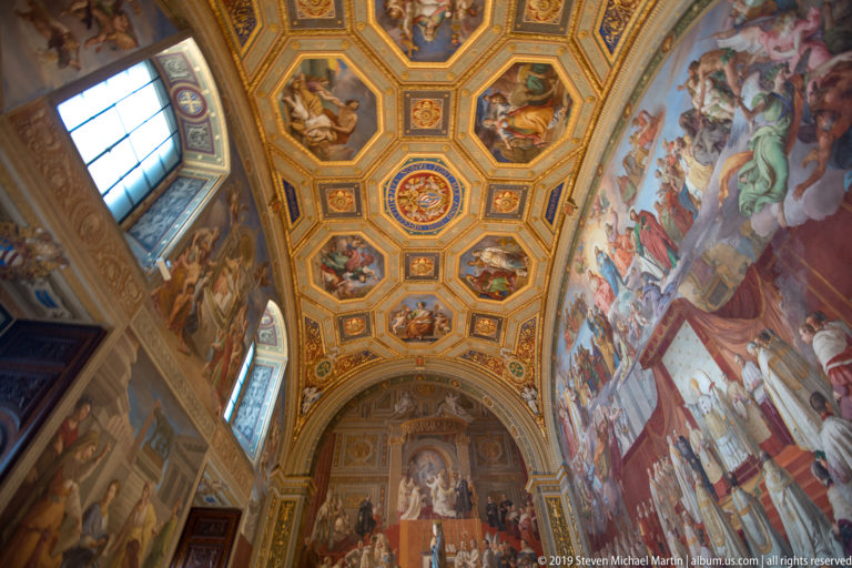 Upward view of a Vatican room with gilded octagonal ceiling panels, tall windows, and monumental wall frescoes.