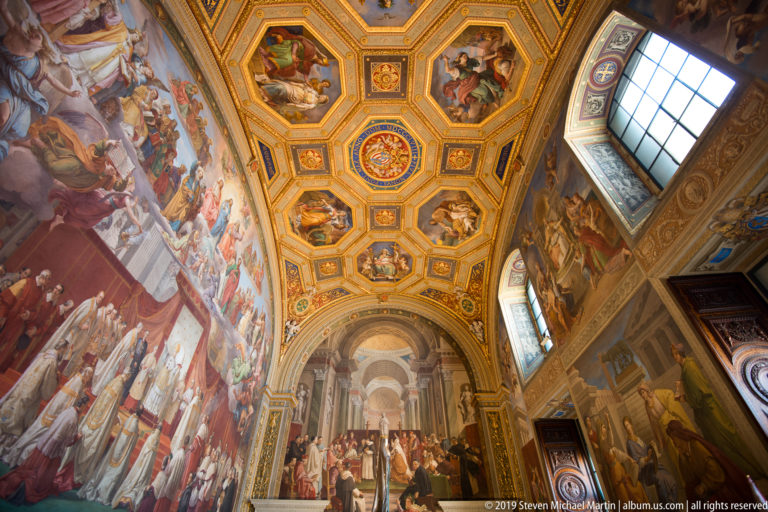 Axial view through a frescoed Vatican room with gilded octagonal ceiling panels, tall windows, and an arched opening ahead.