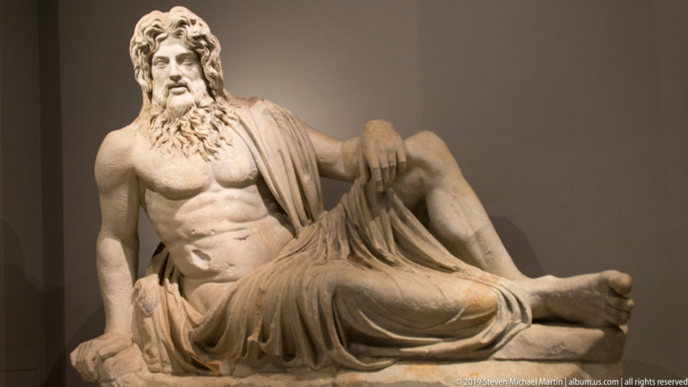 Reclining marble bearded male with muscular bare torso, heavy drapery over his legs, and one knee raised.