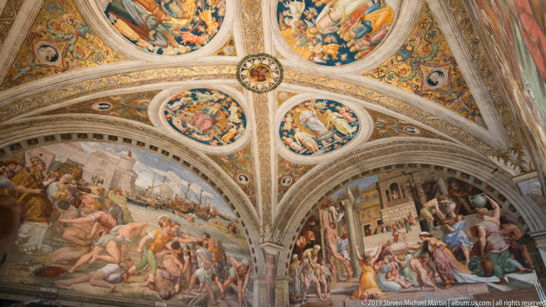 Frescoed groin vault with four circular narrative paintings, gilded ornament, and large crowded wall frescoes below.