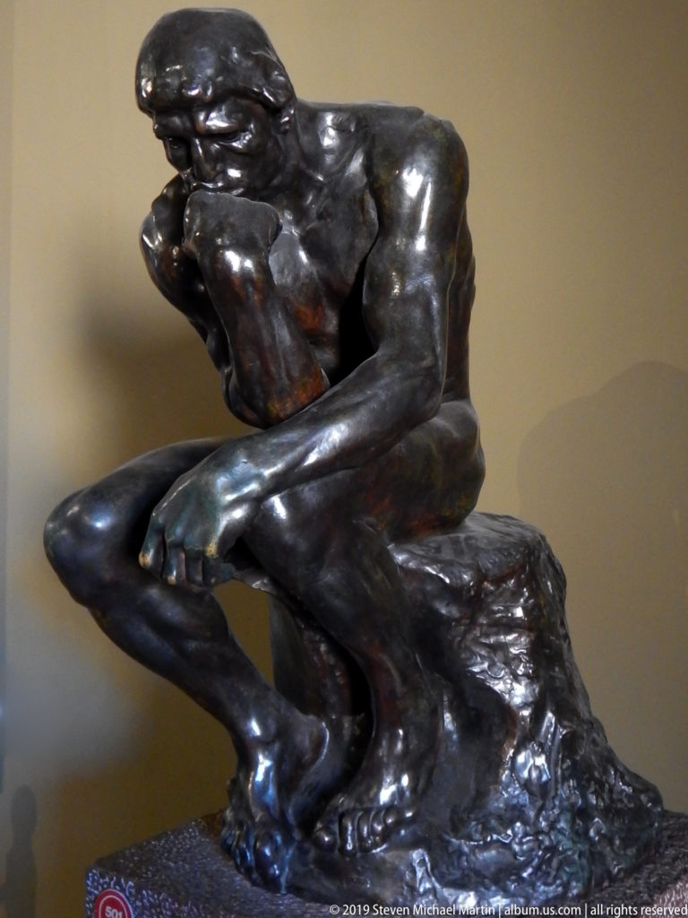 Dark bronze nude male seated in a hunched contemplative pose, head resting on one hand above a rough sculptural base.