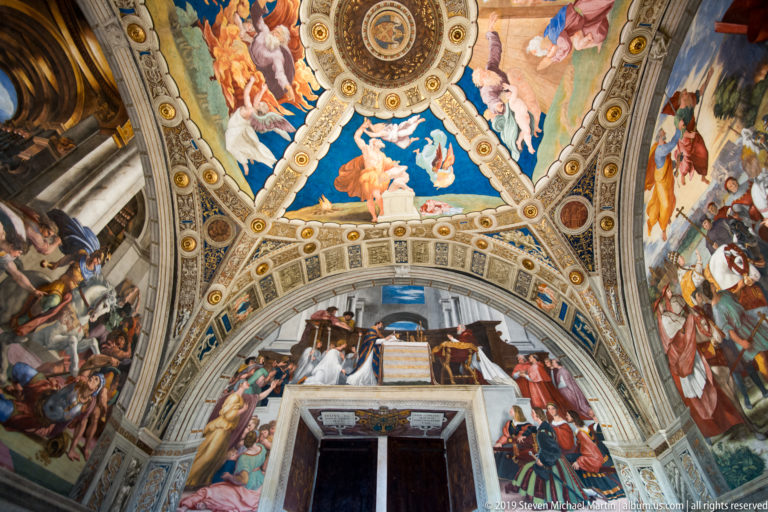 Doorway within a richly frescoed Vatican room, surrounded by large painted figures, blue ceiling fields, and gilded architectural ornament.