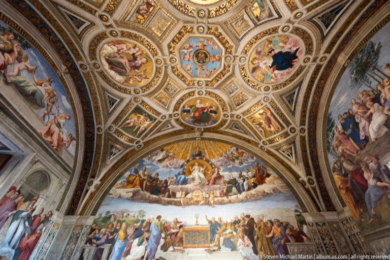Wide view of the Stanza della Segnatura with ornate painted vault, Raphael's Disputa below, and frescoes extending onto adjacent walls.