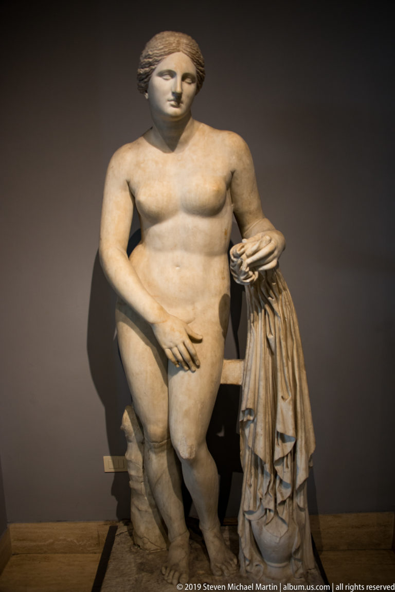 Life-size marble nude female figure standing in contrapposto and holding heavy folded drapery at her side against a dark wall.
