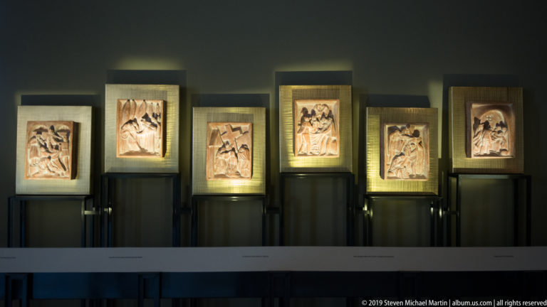 Six carved narrative relief panels displayed in separate illuminated wall cases against a dark gallery interior.