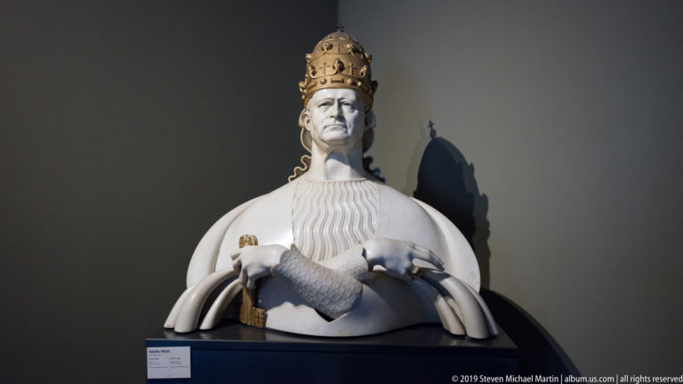 White bust of a pope wearing a papal tiara and patterned vestment, dramatically lit against a dark museum wall.
