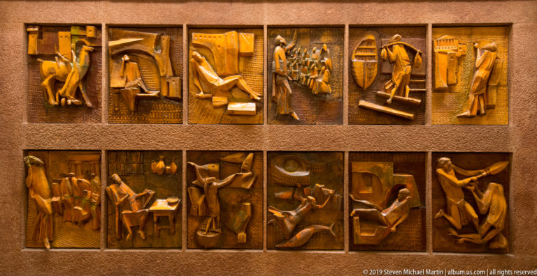 Twelve small bronze relief panels arranged in two rows, each containing stylized human figures in narrative scenes.