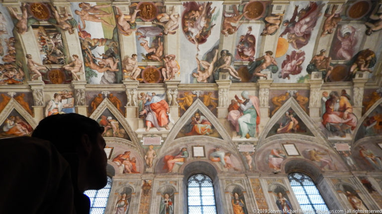 Silhouetted visitor in foreground looking toward the frescoed windows, walls, and ceiling of the Sistine Chapel.
