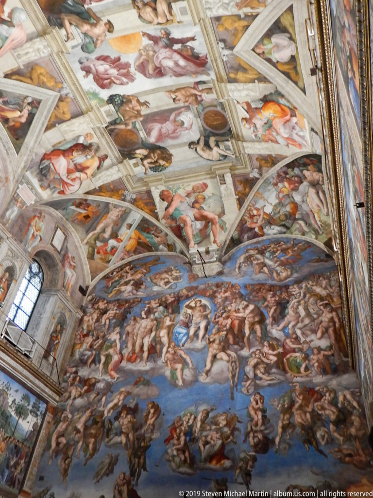 Oblique wide view of Michelangelo's Last Judgment filling the Sistine Chapel altar wall beneath portions of the frescoed ceiling.