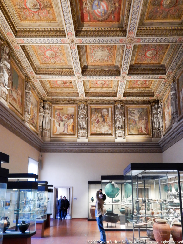 Museum gallery with glass antiquities cases beneath a red-and-gold coffered ceiling and continuous painted figural frieze.