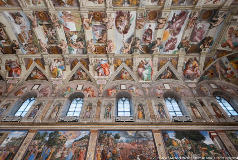 Upward interior view of the Sistine Chapel with tall windows, frescoed wall figures, lunettes, and Michelangelo's ceiling overhead.