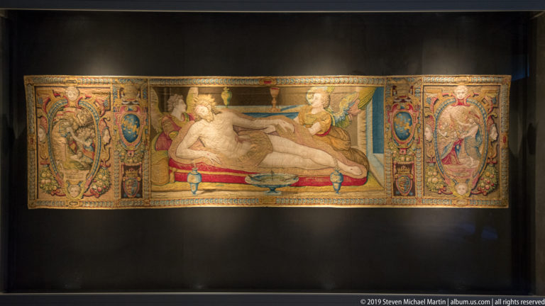 Long tapestry with a reclining central figure, attendants, and richly decorated side borders containing figures, shields, and scrollwork.