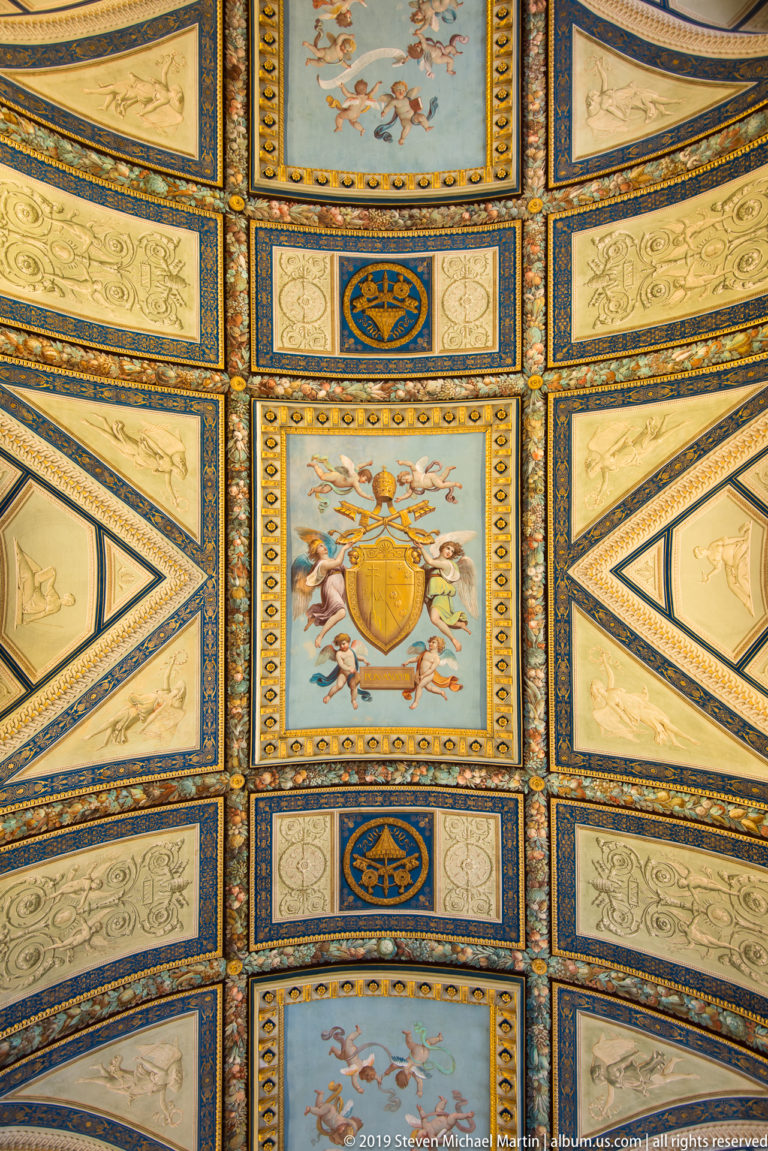 Blue-and-gold geometric ceiling with central papal shield, small figures, grotesques, birds, foliage, and repeated ornamental borders.