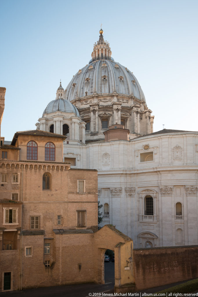 St. Peter's Basilica dome rising behind brick and pale stone Vatican buildings beneath a clear blue sky.